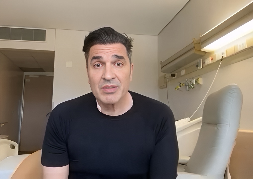 Edu Guedes veste camisa preta e está sentado em uma maca de hospital