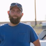 John em um campo de corrida de carros usando chapéu e óculos escuro. Ele está mais magro