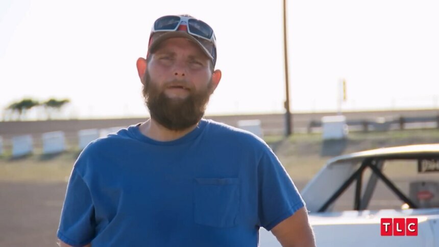 John em um campo de corrida de carros usando chapéu e óculos escuro. Ele está mais magro