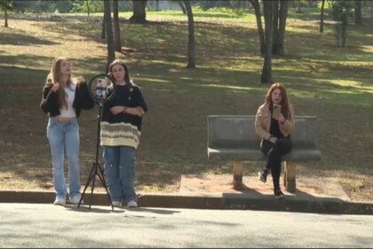 Meninas gravam vídeo em um parque, com um tripé apoiando um celular enquanto outra menina sentada em um banco mexe em seu smartphone em cena do quadro E se fosse Você? do Hoje em Dia