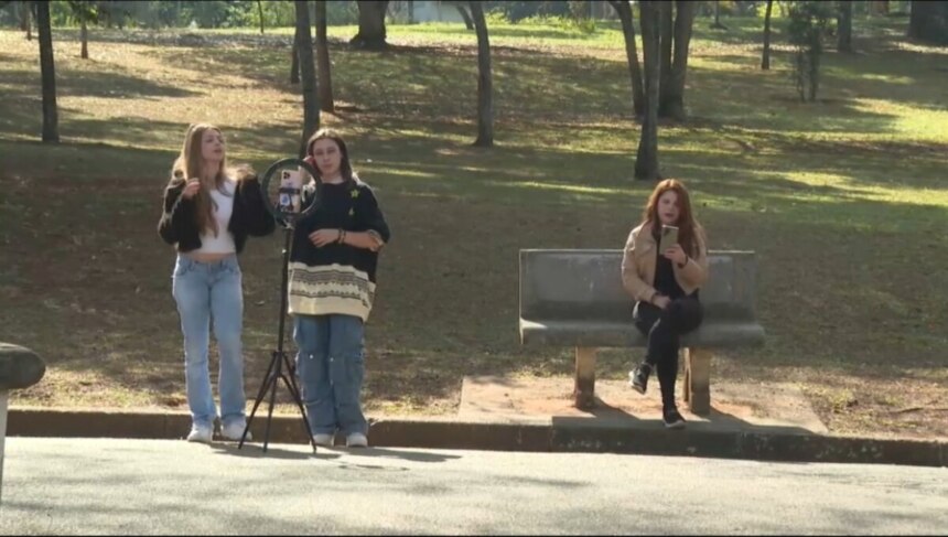 Meninas gravam vídeo em um parque, com um tripé apoiando um celular enquanto outra menina sentada em um banco mexe em seu smartphone em cena do quadro E se fosse Você? do Hoje em Dia