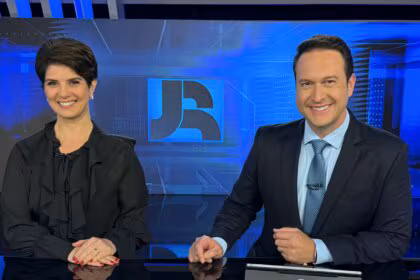 Mariana Godoy e Eduardo Ribeiro sorridentes, sentados na bancada do Jornal da Record, eles vestem roupas pretas