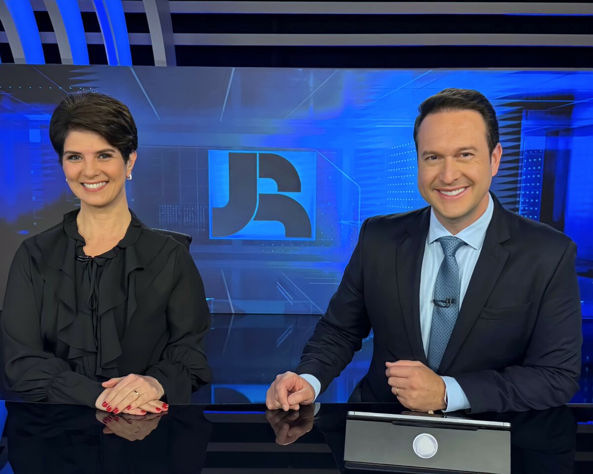 Mariana Godoy e Eduardo Ribeiro sorridentes, sentados na bancada do Jornal da Record, eles vestem roupas pretas
