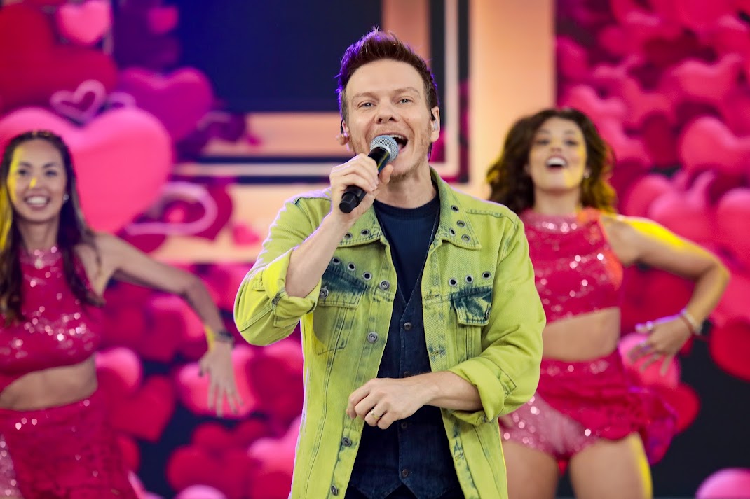 Michel Teló de camisa preta e jaqueta verde, segura microfone e canta no palco do domingo Legal. Por trás dele bailarinas dançam 