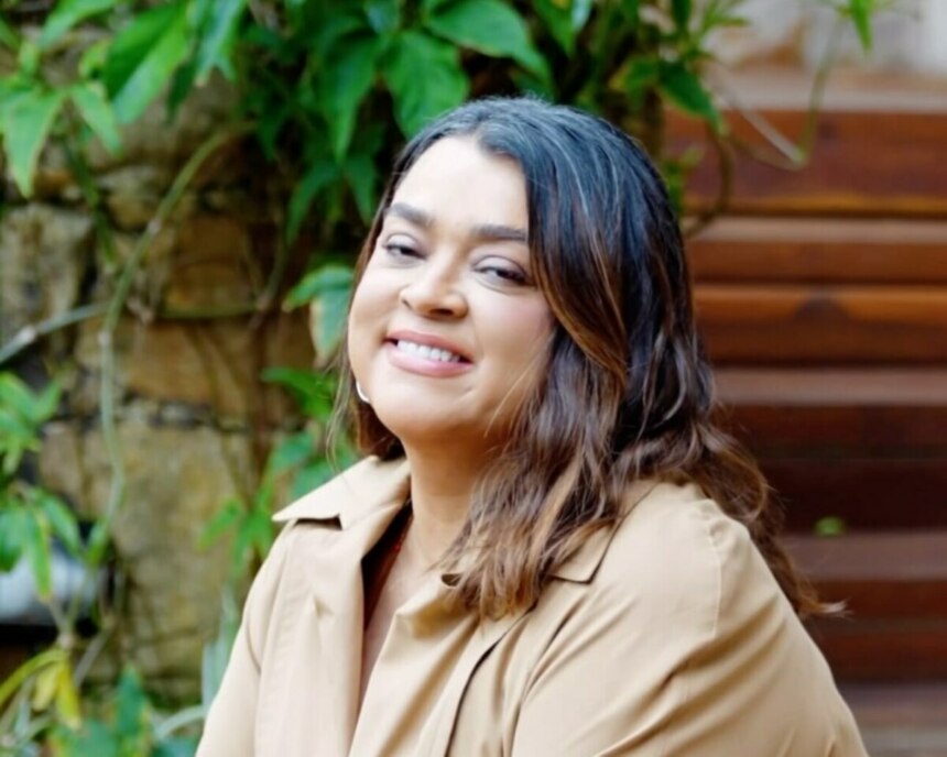 Preta Gil com blusa marrom e sorridente. Ela está sentada em um jardim
