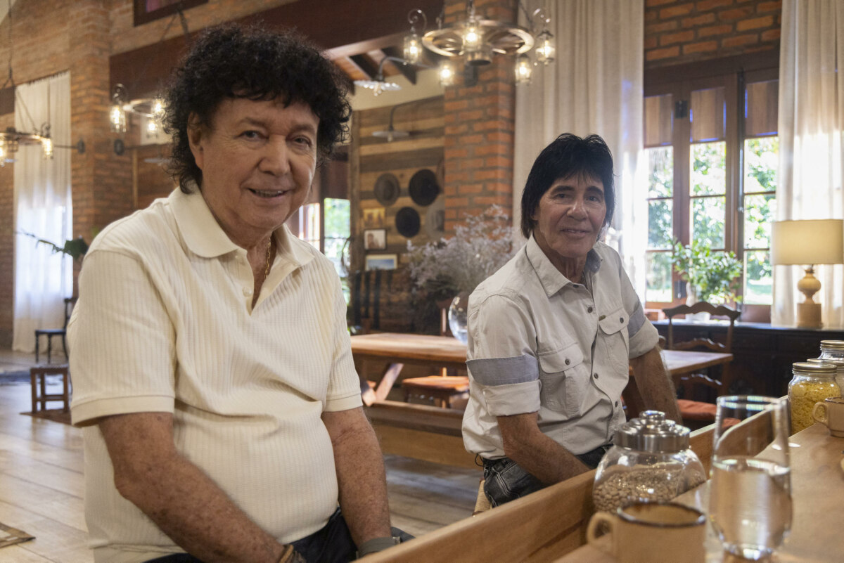 Teodoro e Sampaio sentados na mesa da casa de Daniel no Viver Sertanejo. Eles olham para a câmera e sorriem