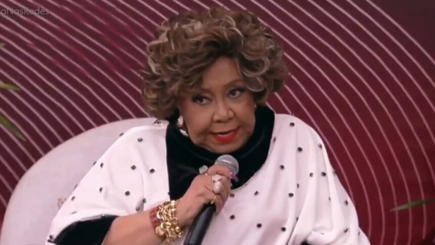 Alcione, com cabelo volumoso e maquiagem destacada, usando um casaco branco com bolinhas pretas e detalhes em veludo preto, segura um microfone e olha diretamente para a câmera. Ela está sentada em um sofá branco, com um fundo desfocado em tons de vermelho e rosa, durante uma participação no programa "É de Casa".