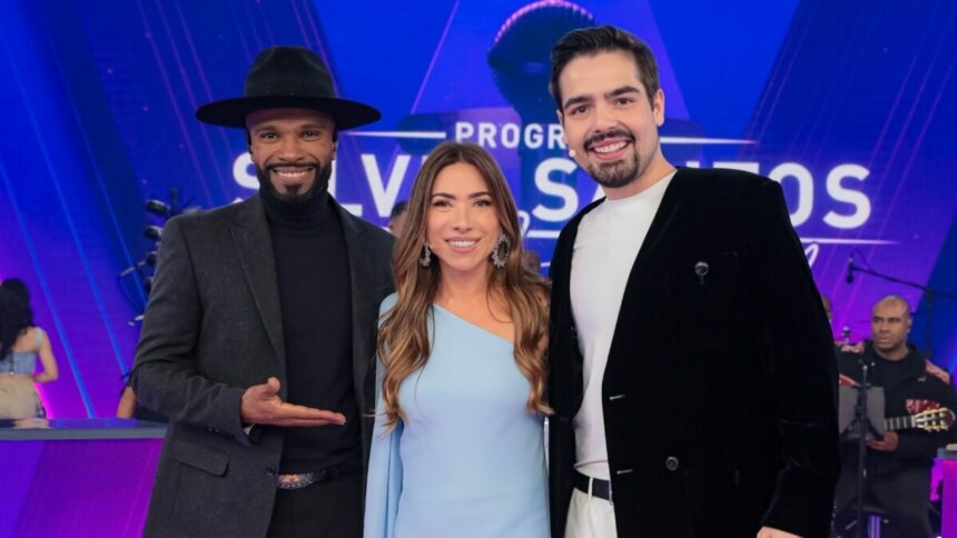 Alexandre Pires (esquerda, de chapéu e roupa escura), Patrícia Abravanel (centro, de vestido azul claro) e João Silva (direita, de blazer preto e camiseta branca) posam sorridentes no palco do "Programa Silvio Santos". Ao fundo, o logo do programa e músicos são visíveis.