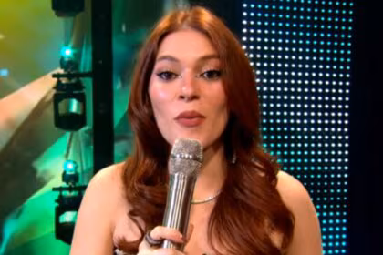 A imagem é um close da apresentadora Ana Clara, sorrindo e falando em um microfone no palco do Estrela da Casa. Ela está usando um vestido brilhante e um colar prateado. O cabelo dela, castanho avermelhado, está solto, no palco do Estrela da Casa