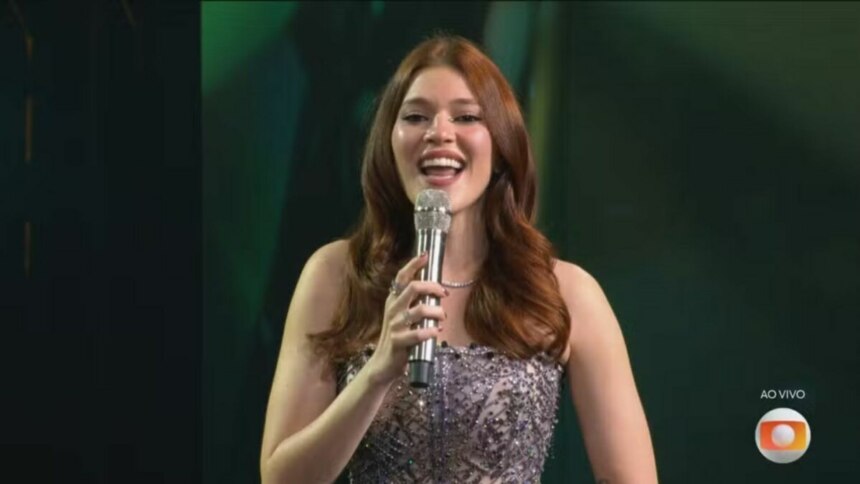 A imagem é um close da apresentadora Ana Clara, sorrindo e falando em um microfone no palco do Estrela da Casa. Ela está usando um vestido brilhante e um colar prateado. O cabelo dela, castanho avermelhado, está solto. Ao fundo, um cenário verde e escuro, com o logo da TV Globo no canto inferior direito.