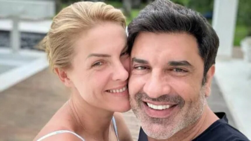 Uma foto de perto de Ana Hickmann e Edu Guedes. Ana, à esquerda, é uma mulher de pele clara com cabelos loiros presos, sorrindo e com a cabeça inclinada para perto de Edu. Edu, à direita, é um homem de pele clara com cabelos escuros e barba por fazer, sorrindo amplamente para a câmera. Ambos parecem estar ao ar livre, com um fundo desfocado.
