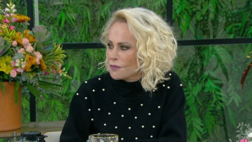 A apresentadora Ana Maria Braga, com um olhar sério, sentada em uma bancada de estúdio de televisão. Ela usa um suéter preto de gola alta com pequenos detalhes brancos. Ao fundo, o cenário é de um ambiente interno com plantas e janelas.