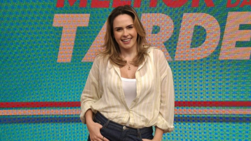 A imagem mostra Ana Paula Renault sorrindo e olhando diretamente para a câmera. Ela usa uma camisa listrada clara sobre uma regata branca e uma calça jeans escura, com as mãos nos bolsos da calça. Ao fundo, um painel digital exibe a frase "MELHOR DA TARDE" em letras grandes e coloridas.