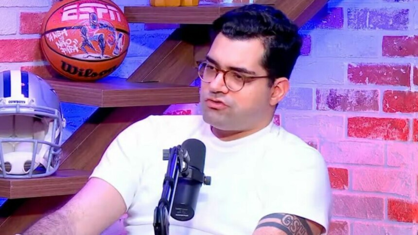 O jornalista Antony Curti em um estúdio de podcast da ESPN. Ele usa óculos e uma camiseta branca, e fala em um microfone com objetos de futebol americano e basquete ao fundo.