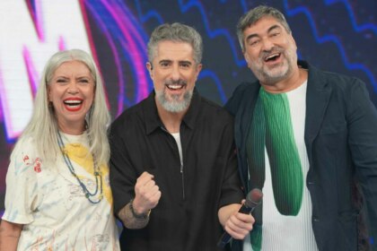 Uma foto de grupo com os apresentadores Astrid Fontenelle, Marcos Mion e Zeca Camargo no cenário do programa 'Caldeirão'. Da esquerda para a direita, estão Astrid de cabelos grisalhos e camiseta estampada, Marcos Mion de camisa preta e Zeca Camargo de jaqueta escura, todos sorrindo para a câmera.
