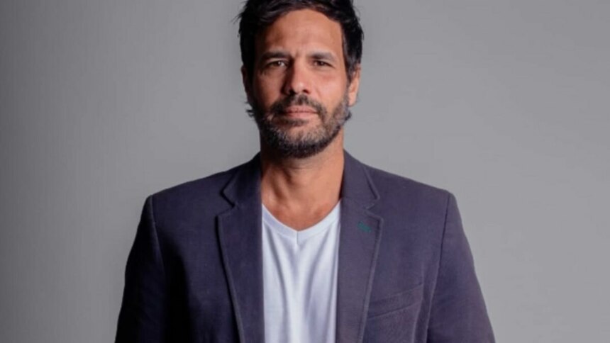 Foto de estúdio do ator Maurício Silveira, com cabelo e barba escuros, vestindo um blazer escuro sobre uma camiseta branca, olhando diretamente para a câmera com uma expressão séria. O fundo é liso e em tom de cinza claro.