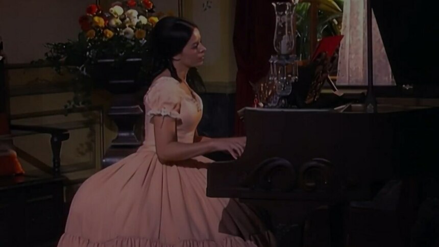 A imagem mostra a atriz Bianca Rinaldi interpretando a personagem "Escrava Isaura". Ela está sentada e toca um piano de madeira escuro. Bianca veste um vestido de época rosa claro, com mangas bufantes. Ao fundo, um vaso de flores, uma cadeira e uma janela escura.