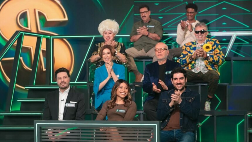 Uma bancada de celebridades em um estúdio de televisão do programa "Show do Milhão". Há nove pessoas, homens e mulheres, sentadas em cadeiras em diferentes níveis. O cenário tem um grande cifrão dourado e luzes verdes. Na primeira fila, o apresentador Danilo Gentili e a apresentadora Blogueirinha, de casaco azul, estão visíveis.