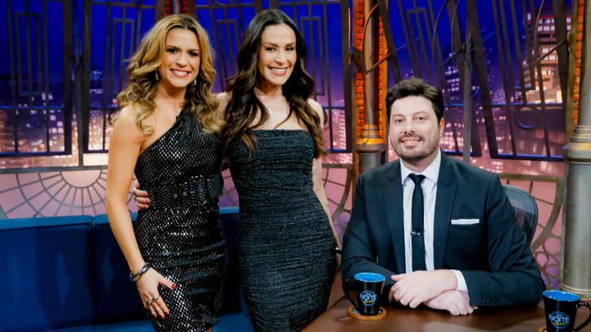 Os apresentadores Danilo Gentili, Beca Milano e Nadja Haddad posam sorrindo juntos no estúdio do programa "The Noite". Danilo está sentado à mesa, e as duas mulheres estão em pé ao seu lado. Beca Milano usa um vestido preto brilhante, e Nadja Haddad usa um vestido preto sem alças.