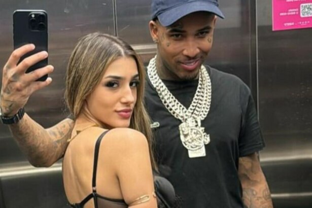 A imagem é uma selfie tirada em um elevador, mostrando Bia Miranda e o cantor Gato Preto. Bia Miranda, à esquerda, usa um top preto e tem os cabelos loiros soltos. Ela olha para trás, em direção à câmera do celular que segura com a mão esquerda. O cantor Gato Preto, à direita, sorri levemente. Ele veste uma camiseta preta e um boné azul-marinho. Ele usa correntes prateadas grossas no pescoço. Ambos têm tatuagens visíveis nos braços. O reflexo do braço de Bia e do celular é visível no espelho do elevador.