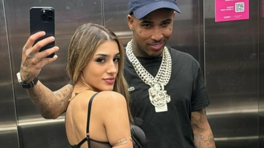 A imagem é uma selfie tirada em um elevador, mostrando Bia Miranda e o cantor Gato Preto. Bia Miranda, à esquerda, usa um top preto e tem os cabelos loiros soltos. Ela olha para trás, em direção à câmera do celular que segura com a mão esquerda. O cantor Gato Preto, à direita, sorri levemente. Ele veste uma camiseta preta e um boné azul-marinho. Ele usa correntes prateadas grossas no pescoço. Ambos têm tatuagens visíveis nos braços. O reflexo do braço de Bia e do celular é visível no espelho do elevador.