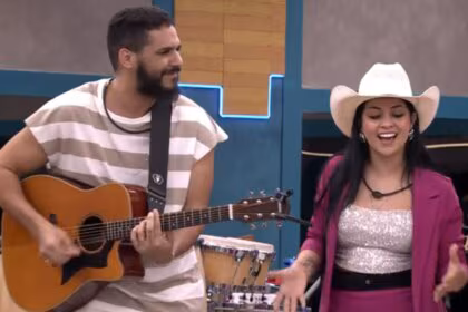 Brenno Casagrande e Biahh Cavalcante no reality Estrela da Casa. Ele toca violão e sorri para ela, que canta usando um chapéu de cowboy e um blazer rosa.