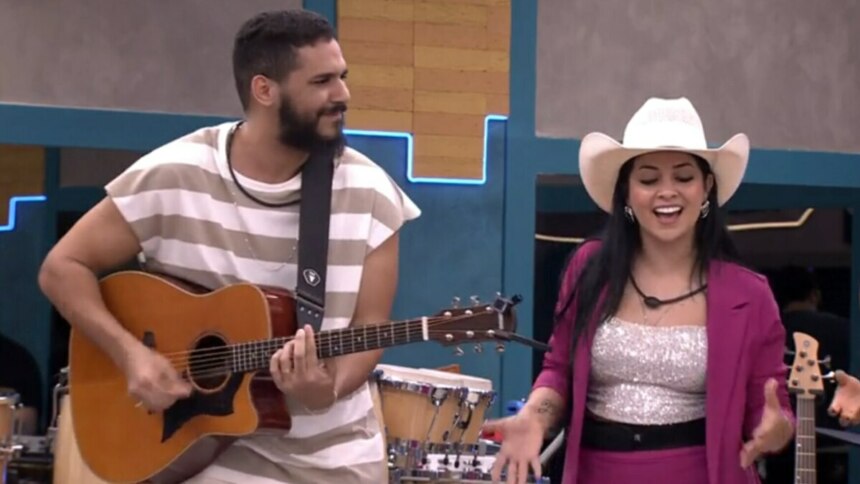Brenno Casagrande e Biahh Cavalcante no reality Estrela da Casa. Ele toca violão e sorri para ela, que canta usando um chapéu de cowboy e um blazer rosa.