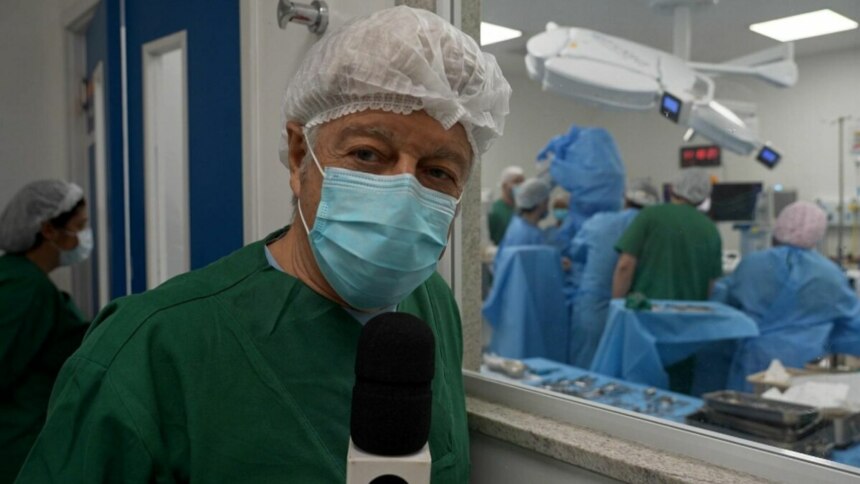 O jornalista Caco Barcellos, em trajes cirúrgicos, máscara e touca, segura um microfone em frente à câmera. Ao fundo, por uma janela de vidro, uma equipe médica trabalha em uma sala de cirurgia, em trecho do Profissão Repórter