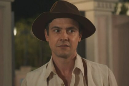 O ator Sérgio Guizé como o personagem Candinho, da novela 'Êta Mundo Melhor!', usa um chapéu de feltro marrom e uma camisa clara, com uma expressão de surpresa ou espanto.
