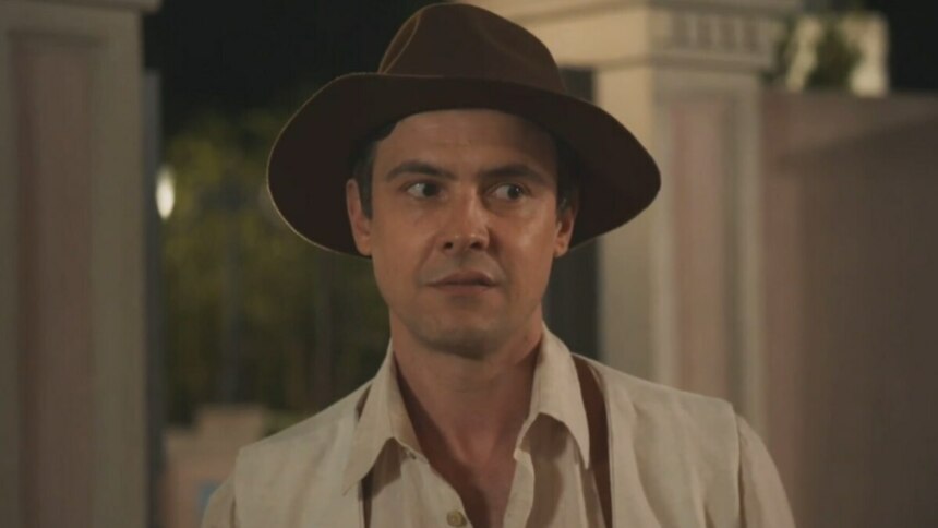 O ator Sérgio Guizé como o personagem Candinho, da novela 'Êta Mundo Melhor!', usa um chapéu de feltro marrom e uma camisa clara, com uma expressão de surpresa ou espanto.