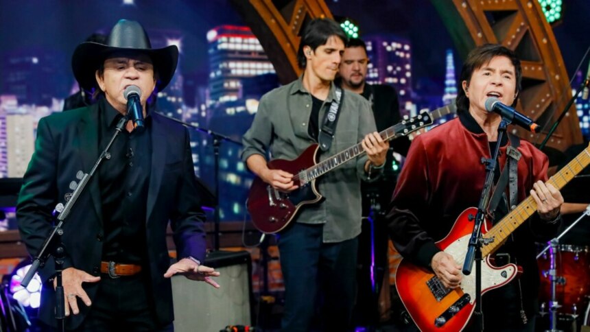 Chitãozinho e Xororó cantando e tocando guitarra no palco do programa The Noite, com a banda ao fundo.