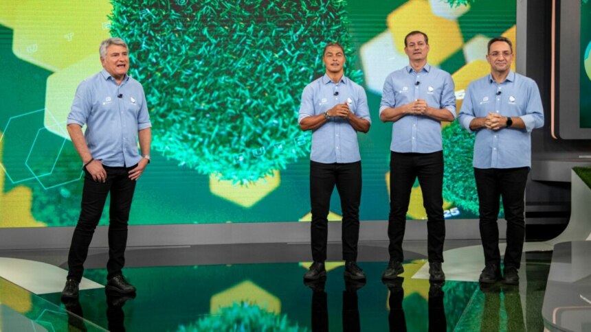 Cleber Machado e sua equipe de comentaristas do Brasileirão na Record posam lado a lado em um estúdio com um fundo temático de gramado e formas geométricas verdes. Da esquerda para a direita: um homem grisalho sorrindo, um homem de camisa polo azul e calça preta com as mãos na frente, Cleber Machado, também de camisa polo azul, e outro comentarista na extrema direita, todos olhando para a frente.