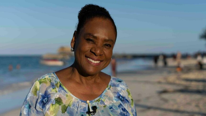 A jornalista Dulcineia Novaes sorri para a câmera em uma praia da República Dominicana, em trecho do Globo Repórter. Ela tem os cabelos presos e usa uma blusa florida de tons azuis e brancos. Ao fundo, um mar azul e areia clara com um barco distante.