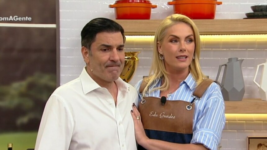 Os apresentadores Edu Guedes e Ana Hickmann em um estúdio de TV, no retorno do quadro "Fica com a Gente". Edu Guedes, de camisa branca, tem uma expressão pensativa. Ana Hickmann, com a mão em seu ombro, o observa. Ela usa um avental com o nome dele bordado.
