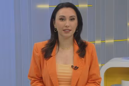 A imagem é um close da apresentadora Eveline Poncio em um estúdio de TV. Ela sorri levemente e olha para a câmera. Eveline usa um blazer laranja sobre uma blusa de cor pêssego, um colar dourado e brincos com formatos de coração. Ao fundo, um cenário claro de televisão e uma caneca com a hashtag "#FazParte" podem ser vistos.
