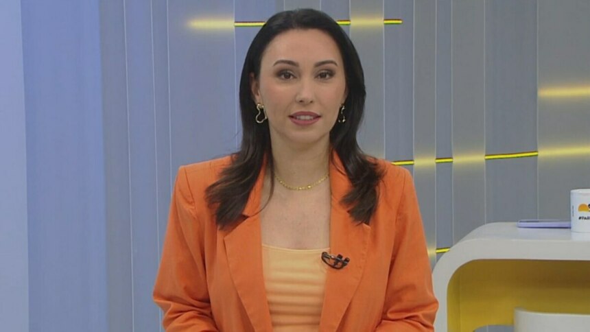 A imagem é um close da apresentadora Eveline Poncio em um estúdio de TV. Ela sorri levemente e olha para a câmera. Eveline usa um blazer laranja sobre uma blusa de cor pêssego, um colar dourado e brincos com formatos de coração. Ao fundo, um cenário claro de televisão e uma caneca com a hashtag "#FazParte" podem ser vistos.