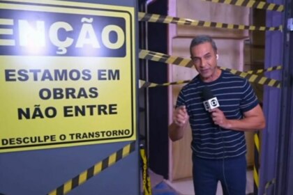 A imagem mostra Fabricio Battaglini em frente a um cenário em obras, possivelmente no programa "Mais Você". Ele segura um microfone com o logotipo da TV Globo e aponta para uma placa de aviso amarela e preta que diz: "ATENÇÃO, ESTAMOS EM OBRAS, NÃO ENTRE" e "DESCULPE O TRANSTORNO". O cenário em volta está com tábuas de madeira e faixas de isolamento amarelas e pretas. O apresentador usa uma camiseta listrada azul e branca e calça jeans.