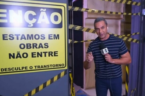 A imagem mostra Fabricio Battaglini em frente a um cenário em obras, possivelmente no programa "Mais Você". Ele segura um microfone com o logotipo da TV Globo e aponta para uma placa de aviso amarela e preta que diz: "ATENÇÃO, ESTAMOS EM OBRAS, NÃO ENTRE" e "DESCULPE O TRANSTORNO". O cenário em volta está com tábuas de madeira e faixas de isolamento amarelas e pretas. O apresentador usa uma camiseta listrada azul e branca e calça jeans.