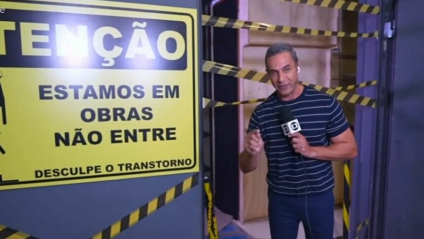 A imagem mostra Fabricio Battaglini em frente a um cenário em obras, possivelmente no programa "Mais Você". Ele segura um microfone com o logotipo da TV Globo e aponta para uma placa de aviso amarela e preta que diz: "ATENÇÃO, ESTAMOS EM OBRAS, NÃO ENTRE" e "DESCULPE O TRANSTORNO". O cenário em volta está com tábuas de madeira e faixas de isolamento amarelas e pretas. O apresentador usa uma camiseta listrada azul e branca e calça jeans.
