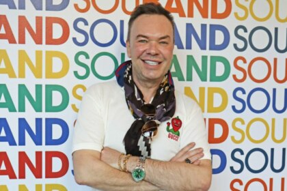 A imagem mostra Felipeh Campos em pé, sorrindo e olhando para a câmera. Ele está de braços cruzados e usa uma camiseta branca com um emblema da Gucci, um lenço no pescoço, pulseiras e um relógio verde. Ao fundo, um painel repete a palavra "BAND" em cores variadas.