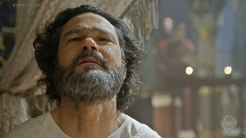 A imagem é um close do ator Gabriel Gracindo em uma cena da novela "O Rico e Lázaro", onde ele interpreta o profeta Daniel. Ele tem o cabelo e a barba grisalha e está olhando para cima com uma expressão séria. Ao fundo, o cenário da novela e uma figura desfocada podem ser vistos.