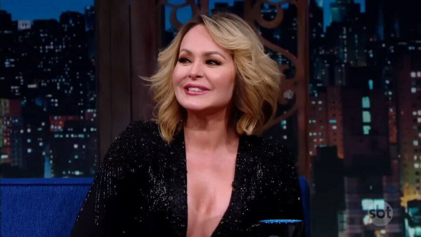 A atriz Gabriela Spanic, com cabelo loiro na altura dos ombros, sentada em um sofá azul em um estúdio de televisão. Cotada para A Fazenda 17, ela está sorrindo e olhando para o lado, vestindo um casaco preto brilhante e decotado. O fundo é um painel digital que simula uma vista noturna de uma cidade, com luzes de prédios.