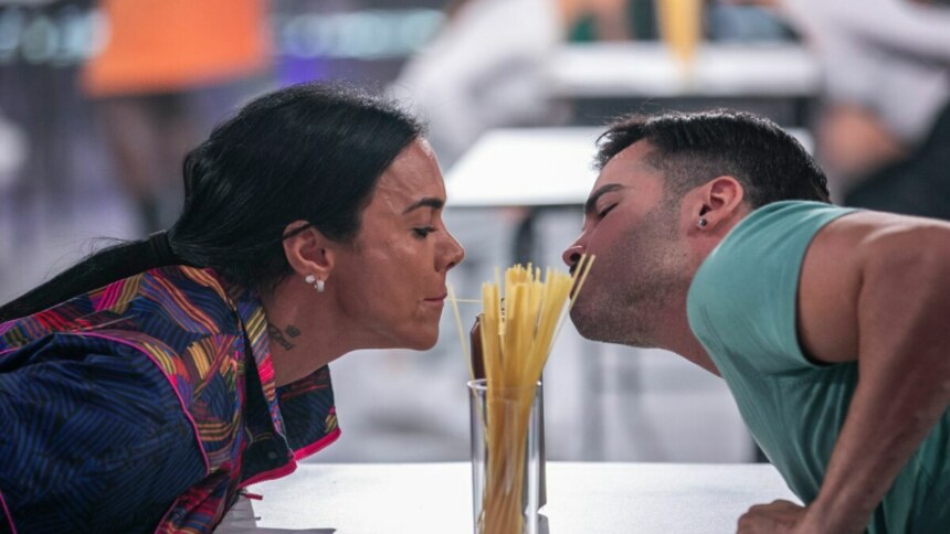 Uma mulher de cabelos escuros presos e um homem com barba e camiseta azul clara se inclinam um em direção ao outro, tentando comer um espaguete comprido que está em um copo alto no centro, durante a "Prova do Macarrão na Lata" no terceiro episódio do "Game dos 100". Ambos estão com a boca perto do macarrão, concentrados na tarefa, em um ambiente de estúdio