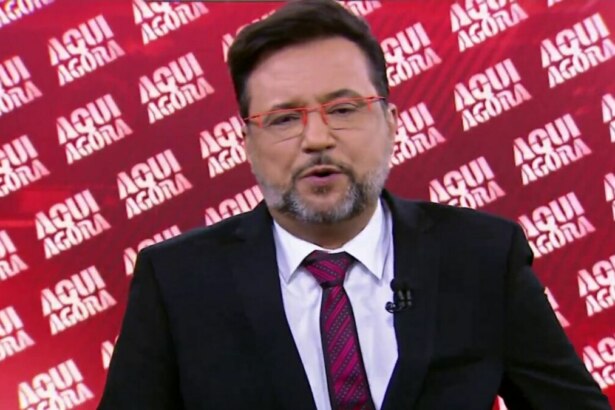 O apresentador Geraldo Luís, com barba e óculos de armação vermelha, em um close-up, olhando para a câmera e falando. Ele veste um terno preto, camisa branca e gravata com listras vermelhas. O fundo é uma parede vermelha com o nome do programa 'AQUI AGORA' escrito repetidamente.