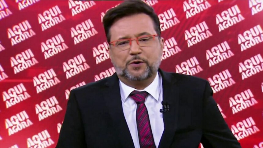 O apresentador Geraldo Luís, com barba e óculos de armação vermelha, em um close-up, olhando para a câmera e falando. Ele veste um terno preto, camisa branca e gravata com listras vermelhas. O fundo é uma parede vermelha com o nome do programa 'AQUI AGORA' escrito repetidamente.