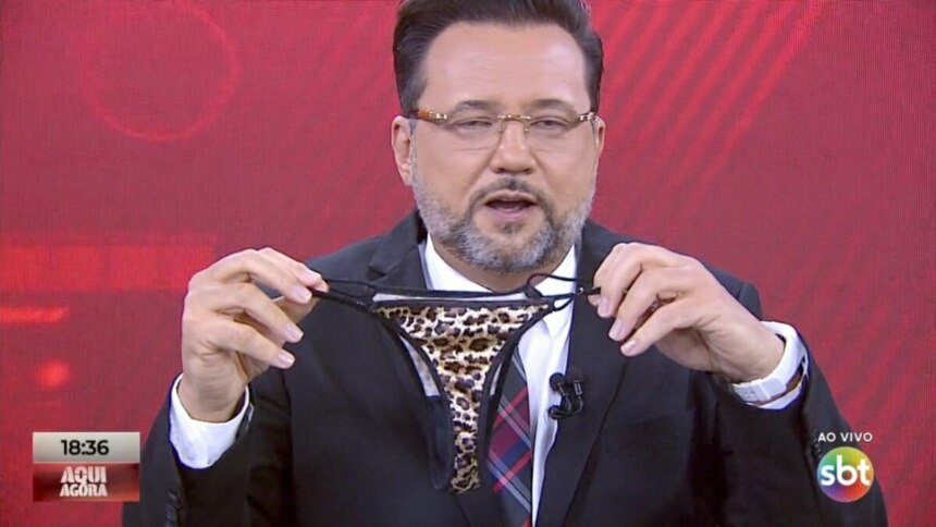 O apresentador Geraldo Luís, usando terno e gravata, segura com as duas mãos uma calcinha de estampa de onça. Ele está no estúdio do programa "Aqui Agora", da emissora SBT, cujo logo e o do programa são visíveis no canto inferior da tela. O fundo é um painel vermelho.