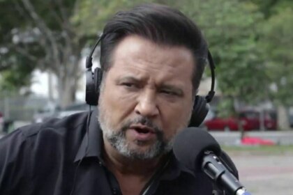 A imagem é um close do jornalista Geraldo Luís durante uma reportagem para o programa "Aqui Agora". Ele está com uma expressão séria, falando em um microfone. Geraldo usa um fone de ouvido preto e uma camisa preta. O fundo está desfocado, mostrando uma rua com árvores e pessoas.