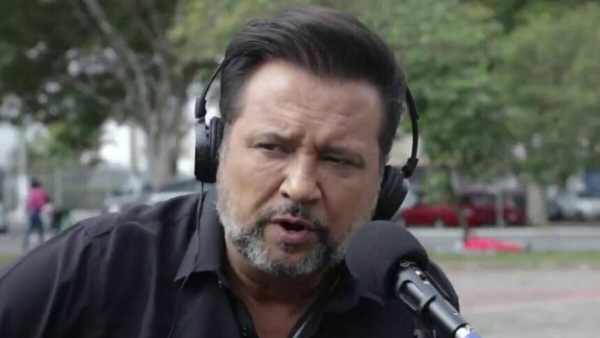 A imagem é um close do jornalista Geraldo Luís durante uma reportagem para o programa "Aqui Agora". Ele está com uma expressão séria, falando em um microfone. Geraldo usa um fone de ouvido preto e uma camisa preta. O fundo está desfocado, mostrando uma rua com árvores e pessoas.