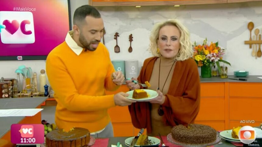 Gil do Vigor e Ana Maria Braga no programa "Mais Você". Ana Maria oferece um pedaço de bolo de cenoura a Gil do Vigor em um prato. Ambos sorriem, e na bancada em frente a eles, há outros bolos. O cenário é a cozinha do estúdio.