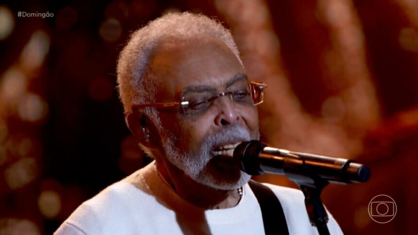 O cantor Gilberto Gil, usando óculos e um suéter branco, canta com os olhos fechados em um microfone durante uma apresentação no palco do Domingão.